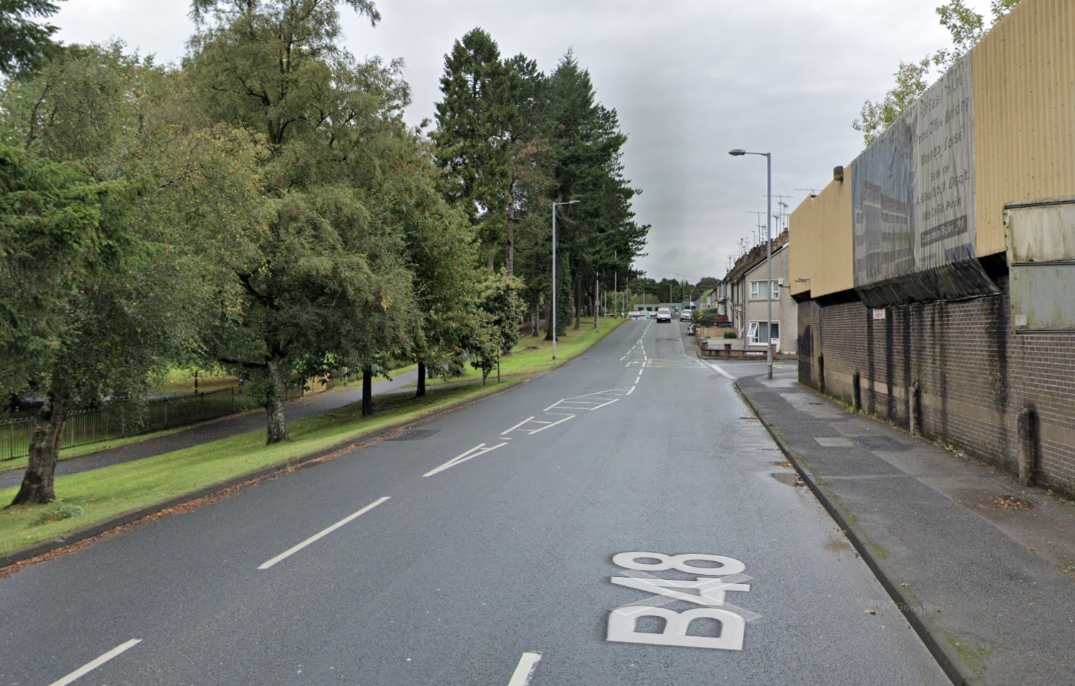 Looking along a two lane road. A high,
yellow metal wall of a police station is on
the right. On the left is a park with a line
of trees beside the road, and footpath running
parallel to the road behind the trees.