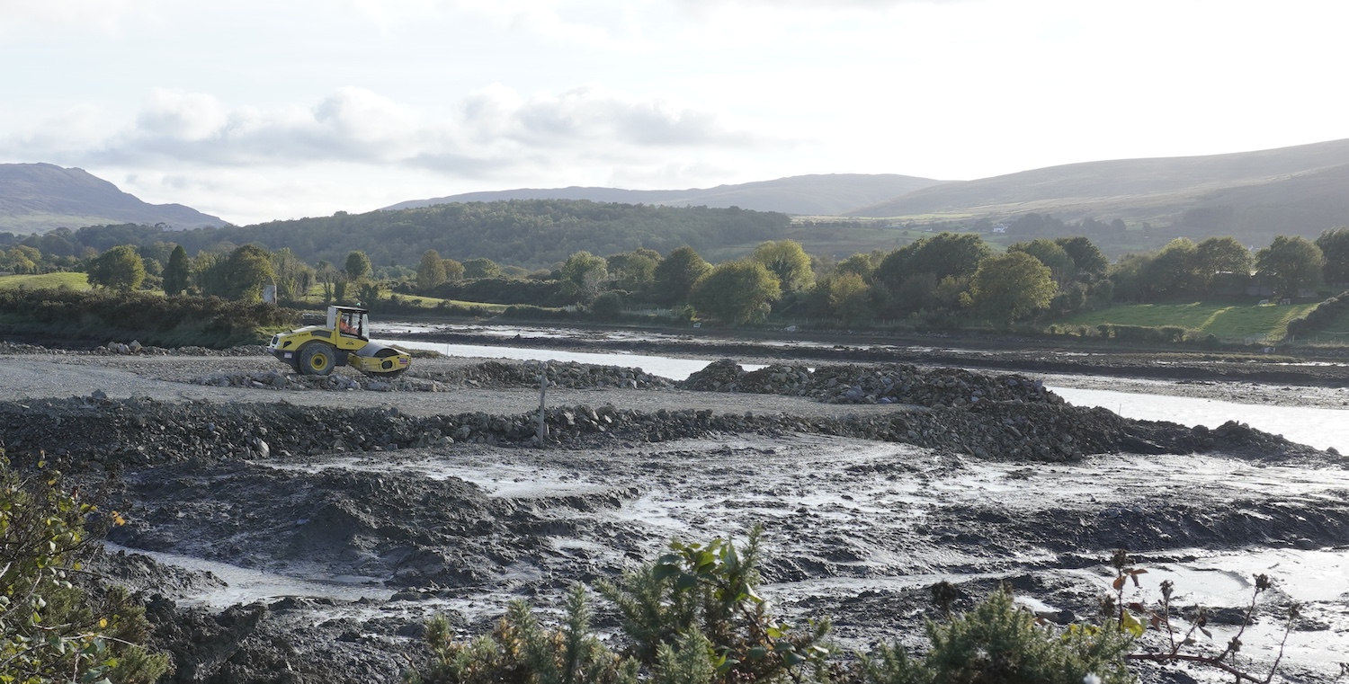 View over water with shore on left. Ahead
is a road roller on some rocks that have been
pushed out onto the mud flats.