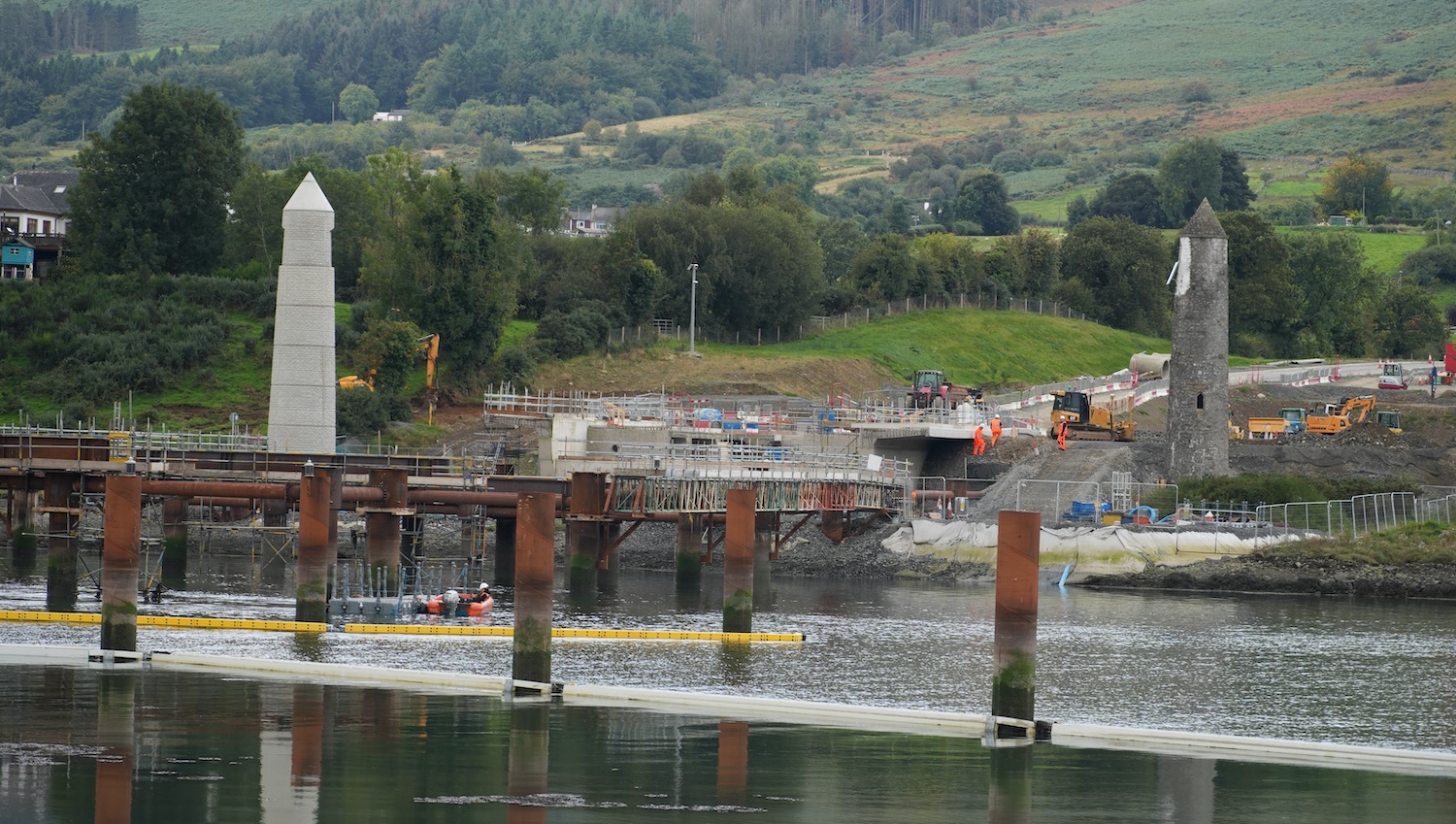 View across the river of a concrete
                          abutment stopping suddently and then a gap
                          where the metal bridge section will go. There
                          are metal posts and frames in the river in the
                          foreground.